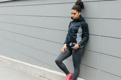 Thoughtful young woman in sports clothing leaning on wall while listening music