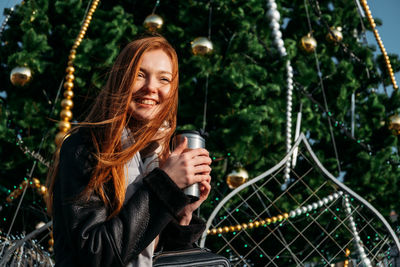Young smiling woman holding coffee cup standing against tree