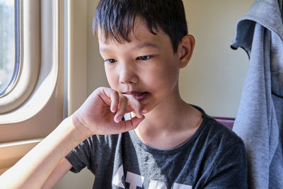 Close-up portrait of young asian thinking boy sitting near window with his hand beside his chin