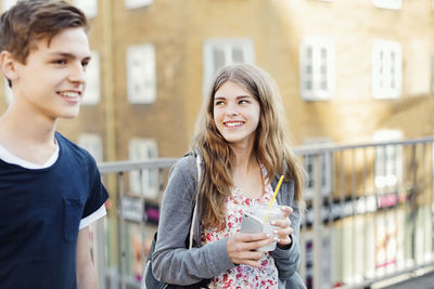 Happy teenager holding disposable glass and smart phone while looking at friend