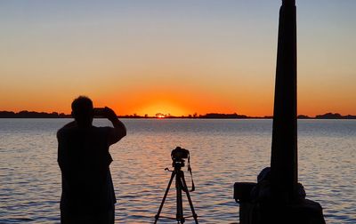 Silhouette photographing by sea against sky during sunset