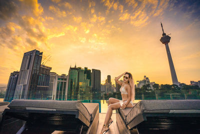 Woman sitting by buildings against sky during sunset