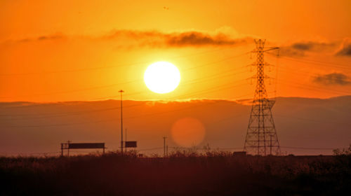 Silhouette electricity pylon on field against orange sky