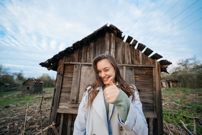 Beautiful girl with long hair in a grey trench coat next to an old wooden house outdoors in spring