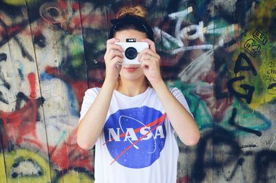Portrait of young woman photographing against graffiti wall