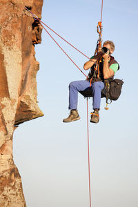 Low angle view of man hanging on rock against sky