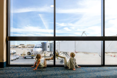 Two girls sitting on floor in airport staring at airplane waiting