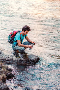 Boy with water bottle crouching at riverbank