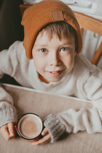 A boy in a sweater and a brown hat drinks cocoa from a red cup. a cozy photo with a mug in hand. 