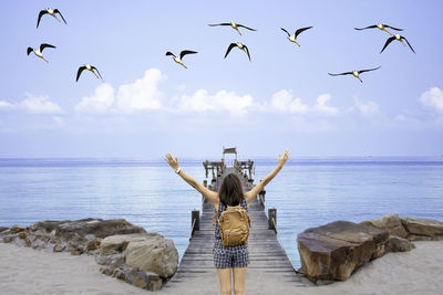 Rear view of woman with arms raised looking at sea while birds flying over beach against sky