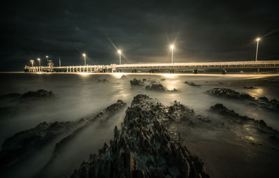 High angle view of rocks against illuminated pier over sea at night