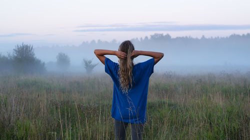 A girlin a blue t-shirt walks through a field with fog at sunset. fog or mist over the field.