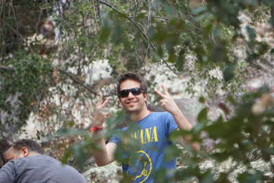 Portrait of man in sunglasses gesturing peace signs amidst trees in forest