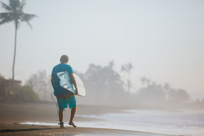 Rear view of man with surfboard walking at beach against sky