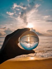 Cropped hand holding crystal ball at beach against sky during sunset