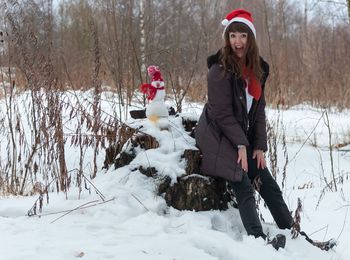 Full length portrait of woman on tree stump at snow covered field