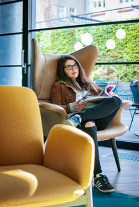 Portrait of young woman reading magazine while sitting on chair against window at home