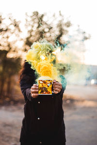 Woman holding mug with yellow emitting smoke against face while standing outdoors