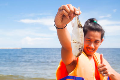 Portrait of man holding fish while gesturing thumbs up against sea