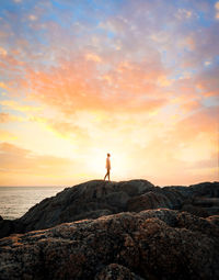 Man standing on rock by sea against sky during sunset