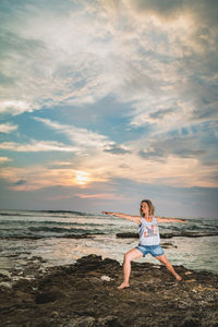 Woman practicing yoga on rocky shore against cloudy sky