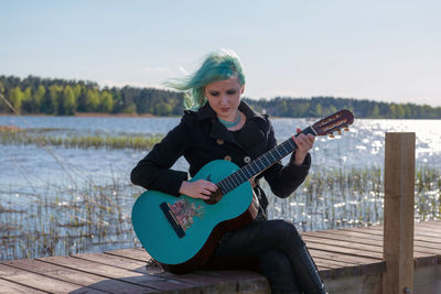 Hipster young woman with turquoise guitar sitting on pier over lake