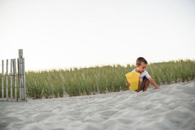 Young boy digging in sand on the beach with a yellow bucket at sunset