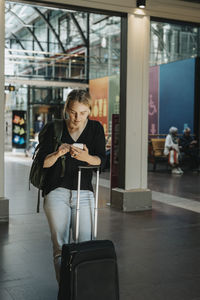 Woman using smart phone while standing with luggage at station