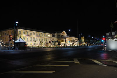 Illuminated street by buildings against sky at night