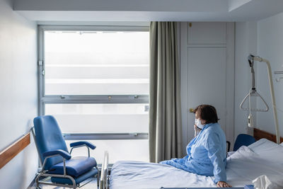 Adult female patient sitting on bed in hospital ward near medicine equipment and talking cellphone