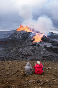 Back view of unrecognizable travelers admiring fagradalsfjall with fire and lava while taking photo and sitting on mount in iceland