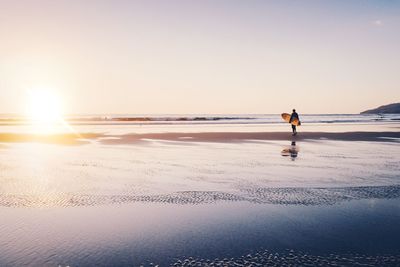 Surfer with surfboard standing at beach against sky during sunset