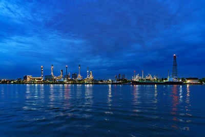 Illuminated factory by sea against blue sky