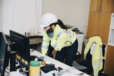 Female engineer wearing hardhat talking on smart phone while leaning on desk in office