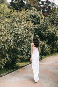 An elegant young woman bride in a wedding dress walks through a green park among plants and trees