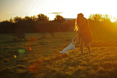 Woman playing with dog on field against sky at sunset