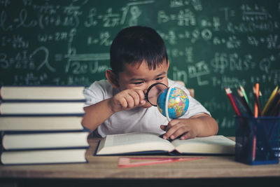 Cute boy studying at table against blackboard