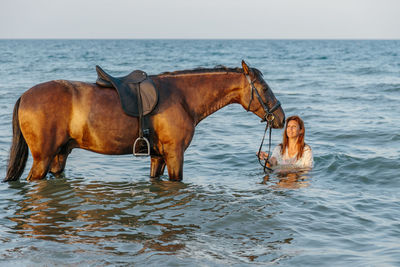 Woman in white dress bathing with her horse in the sea at sunset