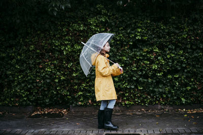 Girl wearing raincoat holding umbrella while walking against leaf wall in city