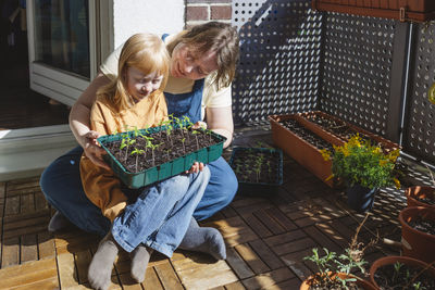 Smiling woman with daughter looking tray of seedlings sitting in balcony on sunny day