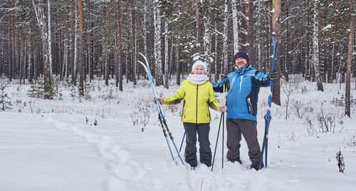 Happy mature couple in winter sportswear standing with cross-country skis in snowy forest, smiling