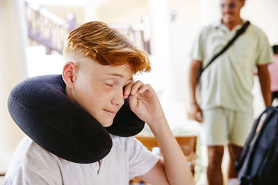 Tired boy with neck pillow rubbing eye in villa during vacation