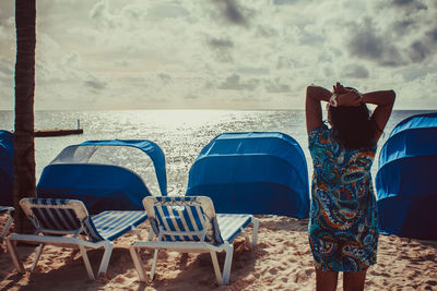 Rear view of woman relaxing on chair at beach against sky