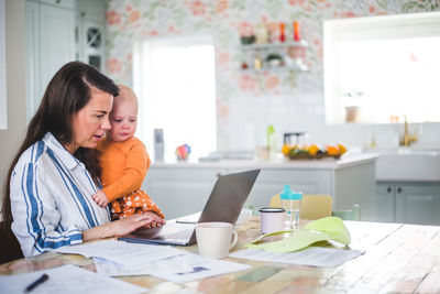 Female entrepreneur carrying loving daughter while using laptop on dining table at home office