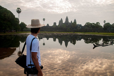 Side view of man looking at angkor wat while standing by lake against sky during sunset