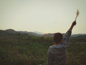 Rear view of woman with hand raised standing on field against sky during sunset