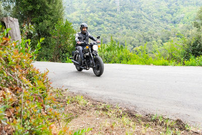 Full body of focused bearded ethnic male biker in black leather jacket and helmet riding modern motorbike on asphalt road amidst lush green trees growing in mountainous valley