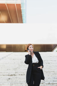 Content female manager in elegant suit walking down the stairs in city and discussing work issues on cellphone while looking away