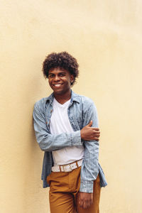 Determined african american male in trendy outfit and with afro hairstyle leaning on stone wall of building and looking at camera