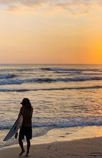 Full length of man with surfboard at beach against sky during sunset